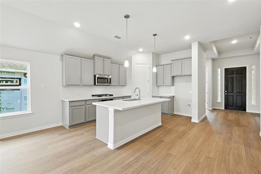 Kitchen with gray cabinets, hanging light fixtures, a kitchen island with sink, light wood-style floors, and stainless steel microwave