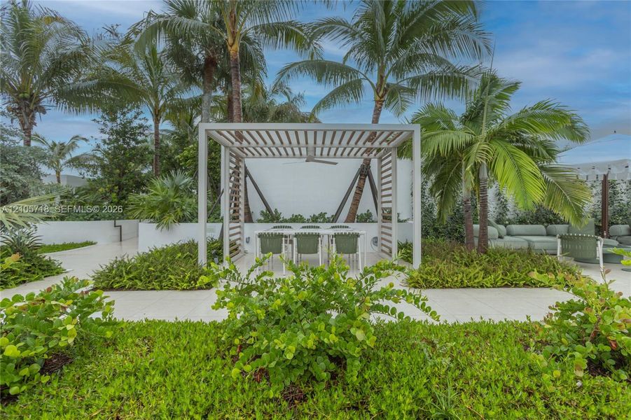 Outdoor patio featuring a white slatted pergola, integrated ceiling fan, light-colored paver flooring, raised garden beds, and mature palm trees