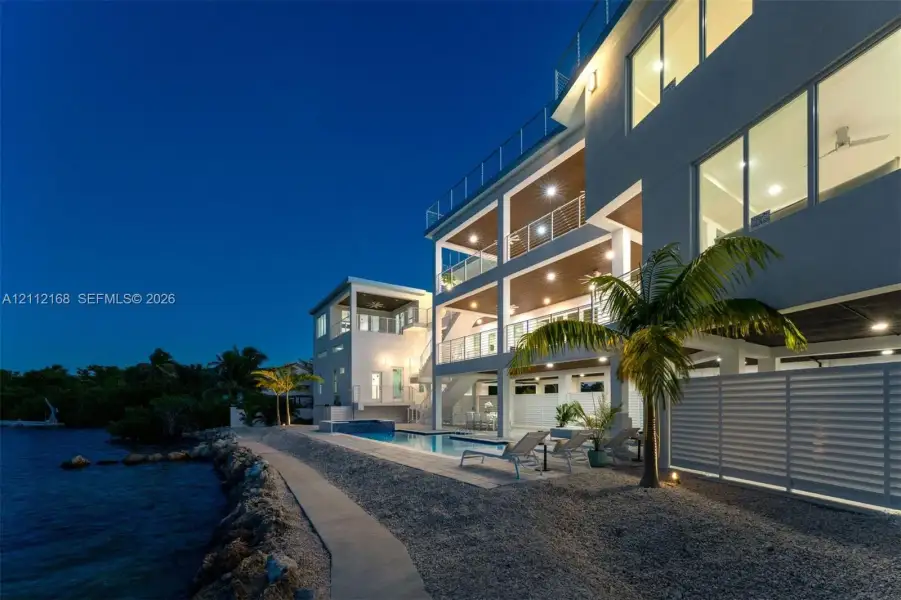 Multi-level exterior featuring stacked balconies with glass railings, a swimming pool with tile surround, and direct waterfront access via a rock-lined shoreline Multi-level exterior featuring stacked balconies with glass railings, a swimming pool with tile surround, and direct waterfront access via a rock-lined shoreline