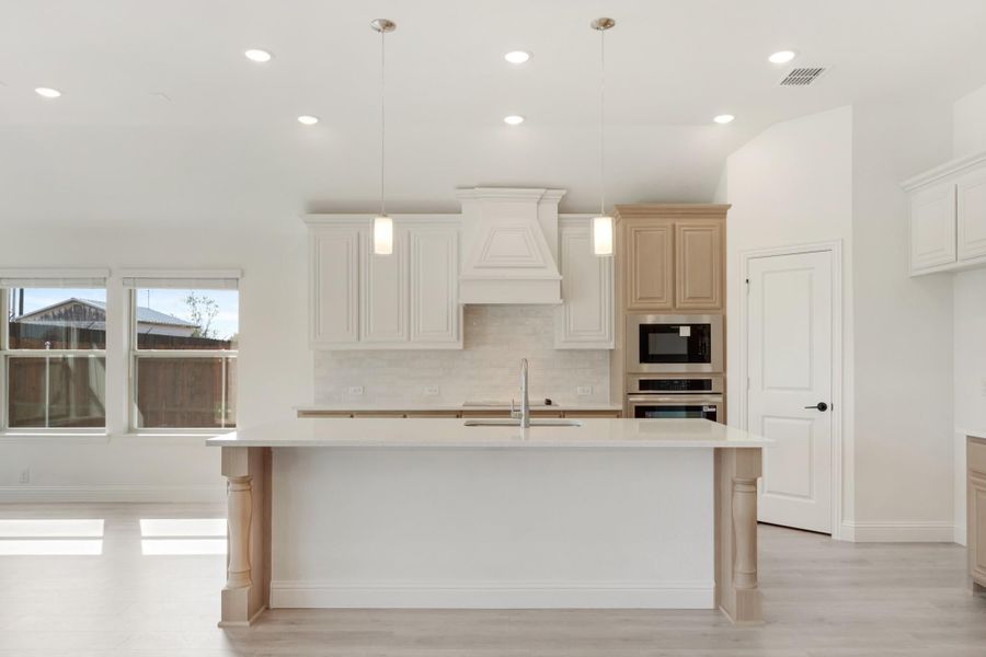 Open kitchen with large white island, pendant lights, custom range hood, and built-in stainless steel appliances