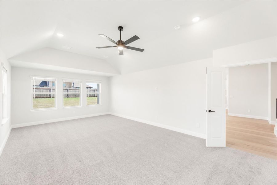 Empty bedroom with vaulted ceiling, ceiling fan, carpet flooring, and three windows overlooking backyard