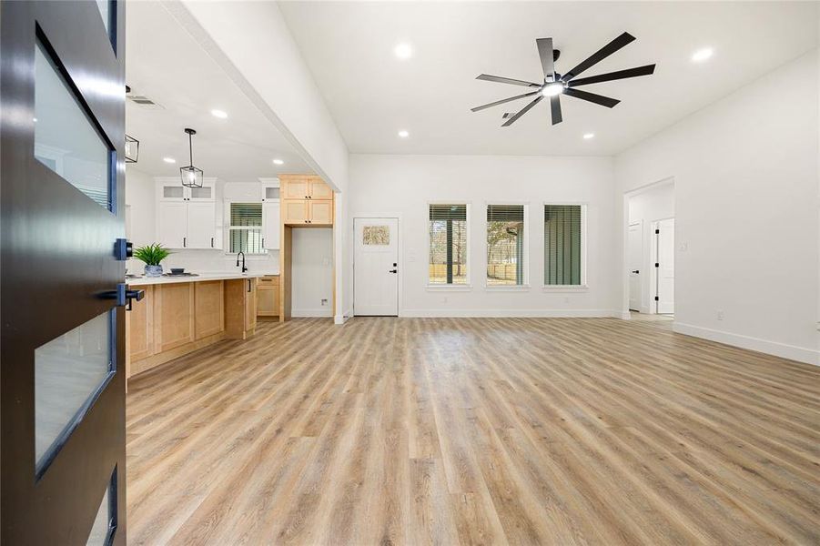 This photo showcases a spacious and modern open-concept living area with sleek wood flooring and ample natural light from large windows. The room features a contemporary ceiling fan and flows seamlessly into a kitchen with light wood cabinetry, adding warmth and functionality.