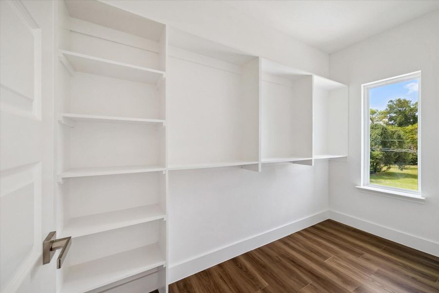A spacious walk-in closet offers excellent organization with extensive built-in shelving, hanging space, and multiple storage sections designed to keep wardrobes and accessories neatly arranged. Crisp white finishes create a clean, bright atmosphere, while warm wood-look flooring adds contrast and continuity with the home’s interior. A large window brings in natural light and overlooks mature greenery, making this thoughtfully designed closet feel open, functional, and inviting. A spacious walk-in closet offers excellent organization with extensive built-in shelving, hanging space, and multiple storage sections designed to keep wardrobes and accessories neatly arranged. Crisp white finishes create a clean, bright atmosphere, while warm wood-look flooring adds contrast and continuity with the home’s interior. A large window brings in natural light and overlooks mature greenery, making this thoughtfully designed closet feel open, functional, and inviting.
