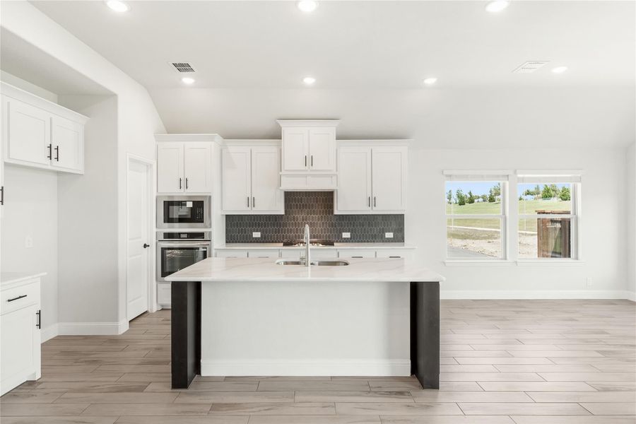 Kitchen with white cabinets, center island with sink, dark tile backsplash, and stainless steel appliances on light wood floors.