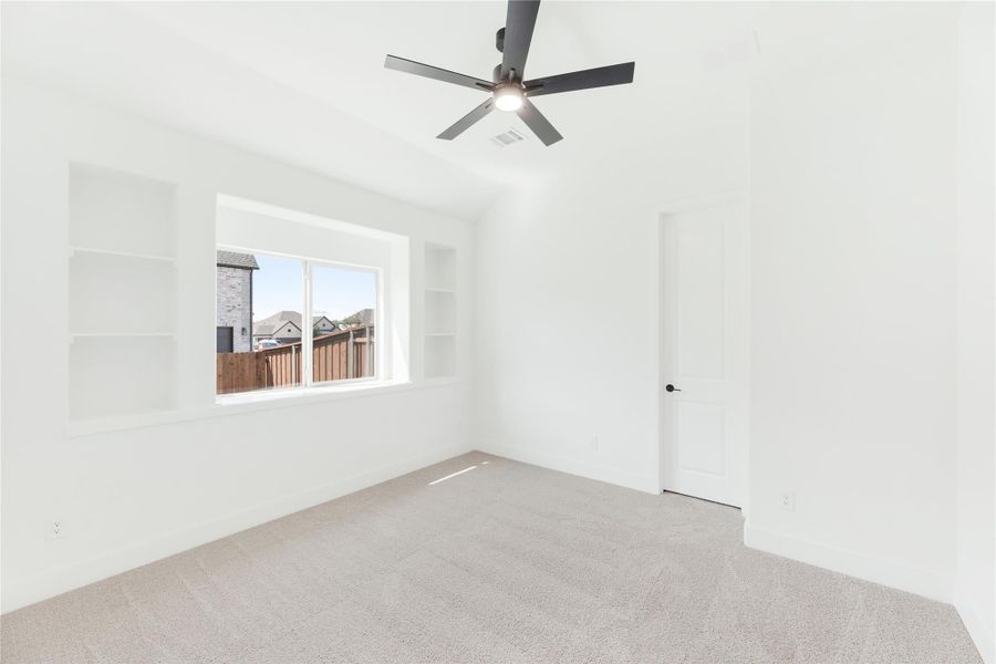 Empty bedroom with carpet, ceiling fan, built-in wall shelves, and a window overlooking a backyard fence.