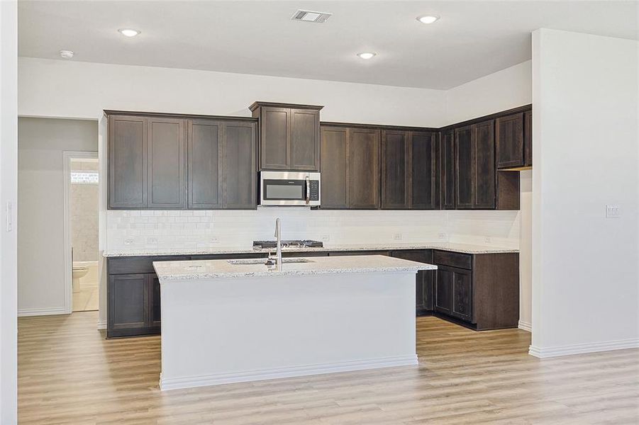 Kitchen with light stone counters, dark brown cabinetry, an island with sink, and light hardwood / wood-style flooring