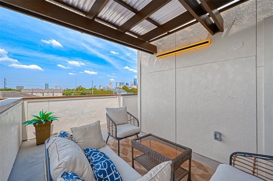 This photo showcases a cozy outdoor patio with modern seating, a stylish coffee table, and a partial city view. The space is covered with a wooden pergola and features a warm, ambient light, creating a perfect spot for relaxation or entertaining.