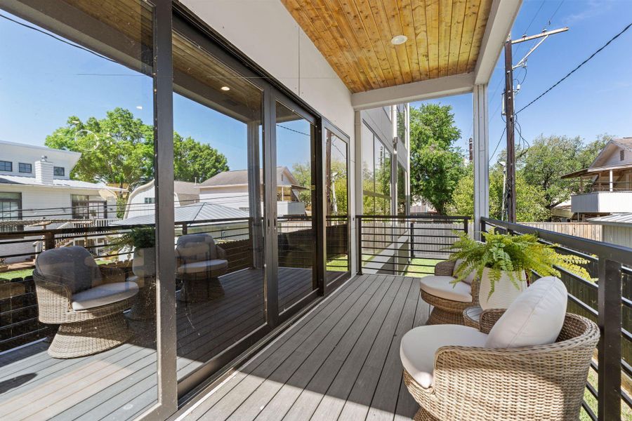 The covered back patio is finished with a tongue-and-groove cedarwood ceiling and recessed lighting — the same material warmth that runs through the porte-cochere and upper balcony. Sliding glass doors connect directly to the interior and black railings frame the yard view below.