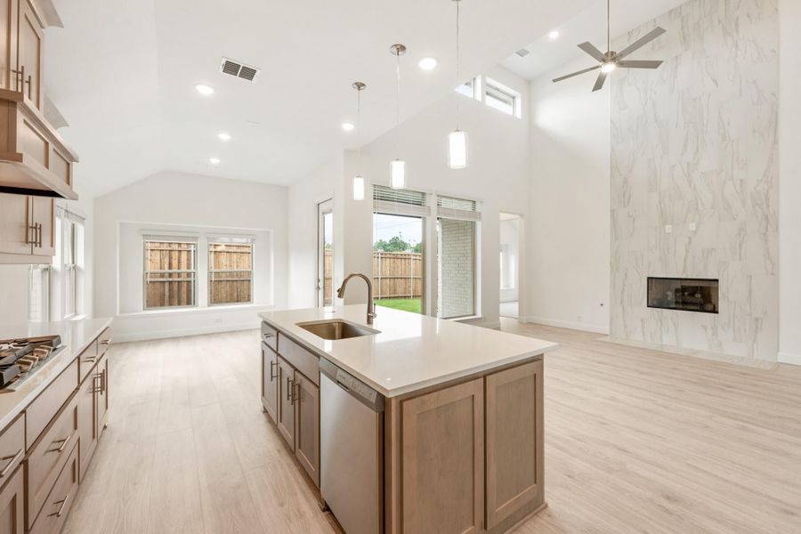 Open-concept kitchen with large island, sink, wood cabinets, high vaulted ceiling, and marble fireplace wall adjoining living area.
