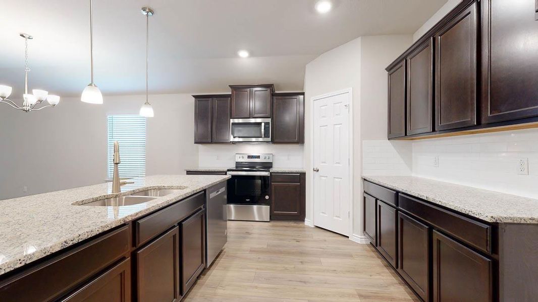 Kitchen featuring dark wood finish cabinetry, stainless steel appliances, light stone counters, light wood-style floors, and suspended lighting