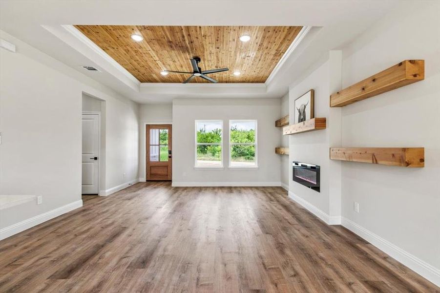 Spacious living area featuring wood-finish flooring, a tray ceiling with wood paneling and recessed lighting, and a modern fireplace with built-in wooden shelving