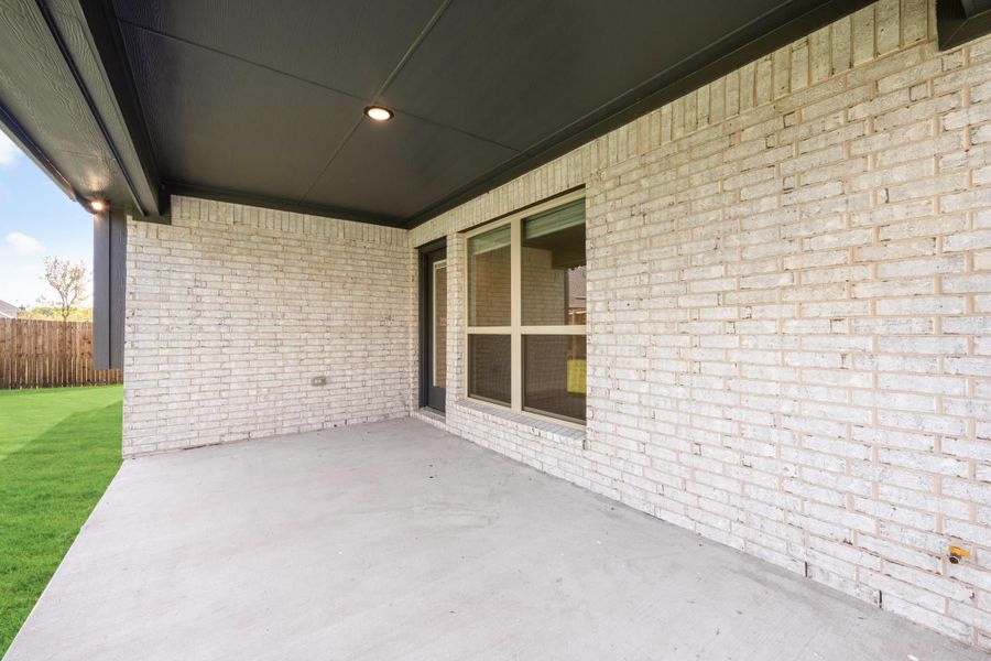 Covered back patio with concrete floor, light brick exterior, sliding window, and fenced green backyard.