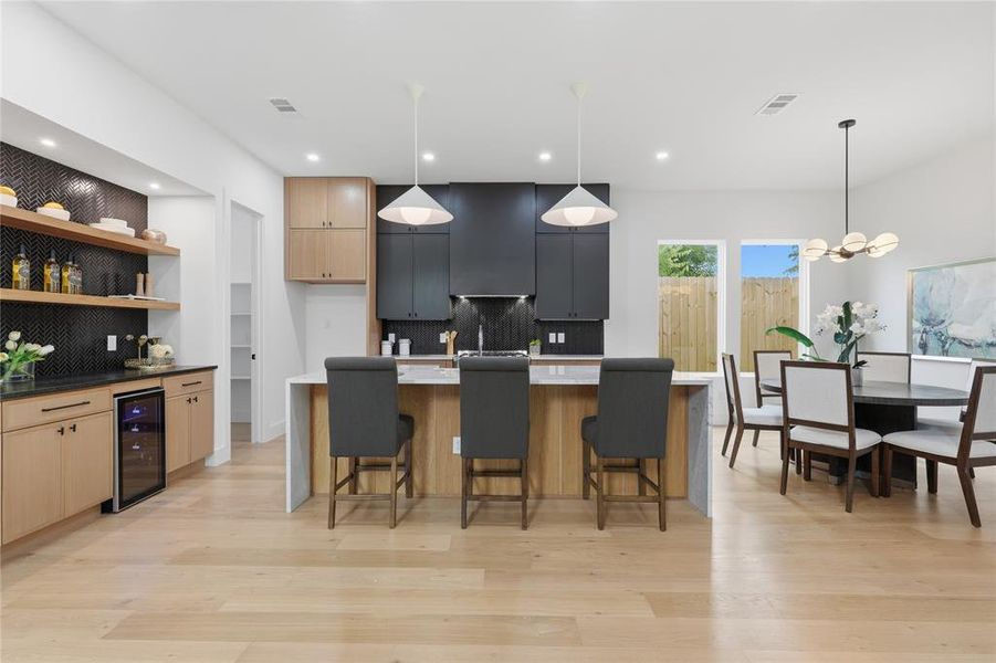 Open-concept kitchen and dining area featuring light wood-finish flooring, a kitchen island with seating, a built-in beverage refrigerator, a black tile backsplash, and contemporary pendant lighting Open-concept kitchen and dining area featuring light wood-finish flooring, a kitchen island with seating, a built-in beverage refrigerator, a black tile backsplash, and contemporary pendant lighting