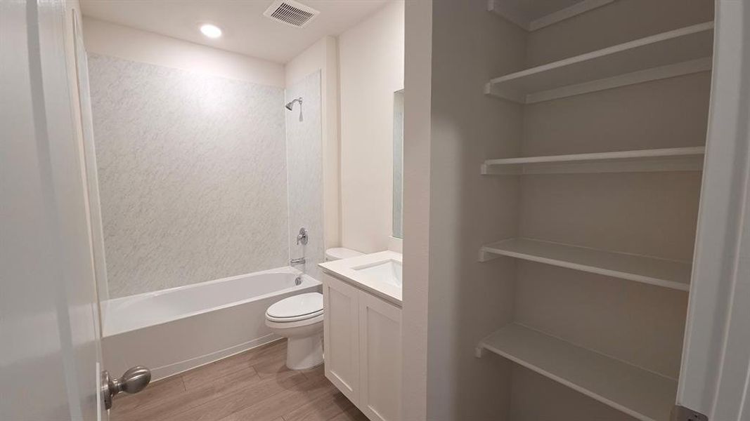 Full bathroom featuring a white tub with a textured shower surround, wood-finish flooring, a white vanity with an integrated sink, and an adjacent built-in linen closet with multiple shelves