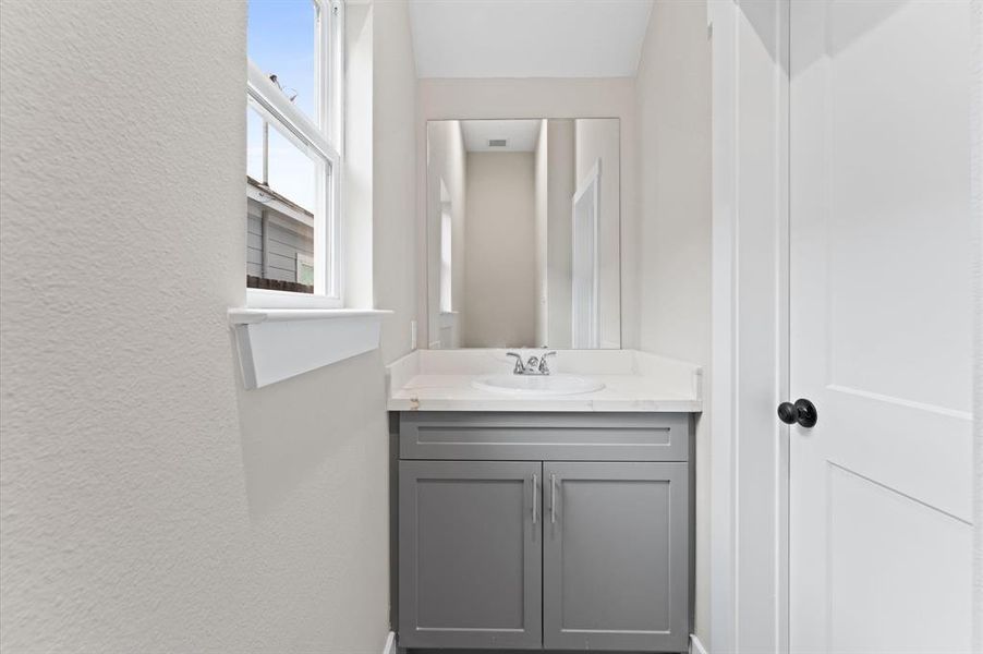 This is a compact, modern bathroom featuring a sleek gray vanity with a quartz countertop, a large mirror, and bright natural light from a side window.