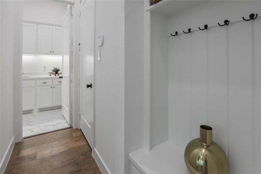 Entryway featuring wood-finish flooring, white paneling with coat hooks, and a laundry room beyond with white cabinetry, under-cabinet lighting, and stone flooring