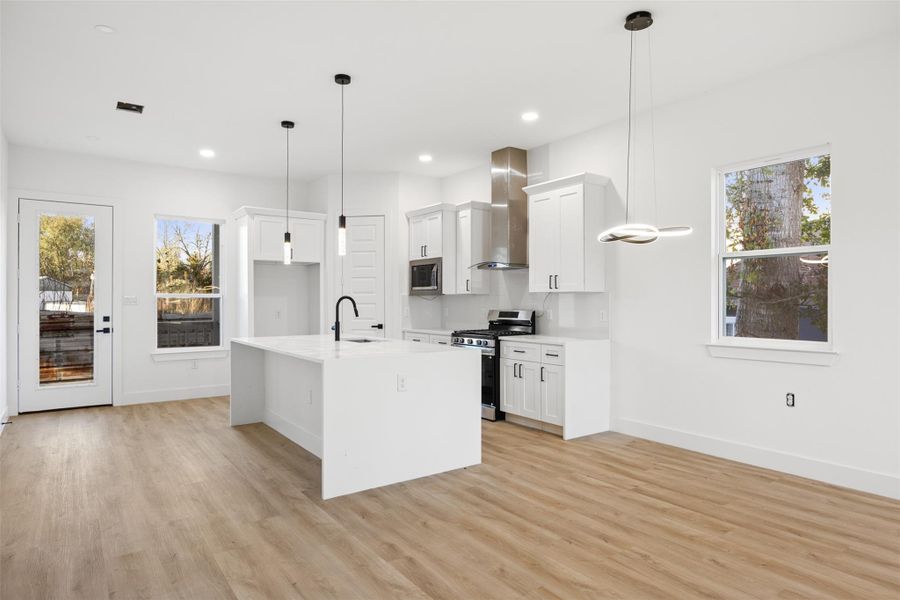 Bright and modern kitchen with sleek white cabinetry, stainless steel appliances, and a spacious island. Features wood-like flooring, pendant lighting, and ample natural light from large windows and glass door.