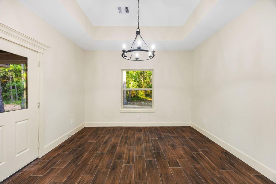 The dining room features rich wood-look flooring, a stylish modern chandelier, and a large window offering natural light and views of the greenery outside. The neutral walls and recessed ceiling add a spacious and airy feel.