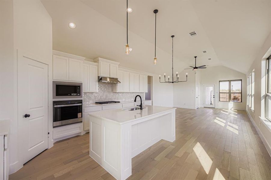Kitchen featuring white cabinetry, stainless steel appliances, hanging light fixtures, a center island with sink, and light wood finished floors Kitchen featuring white cabinetry, stainless steel appliances, hanging light fixtures, a center island with sink, and light wood finished floors