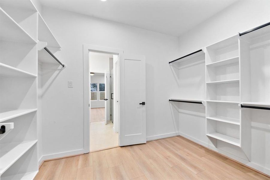 This photo showcases a spacious walk-in closet with ample shelving and hanging space. It features light wood flooring, white walls, and a door leading to another room, offering a bright and organized storage solution. This photo showcases a spacious walk-in closet with ample shelving and hanging space. It features light wood flooring, white walls, and a door leading to another room, offering a bright and organized storage solution.