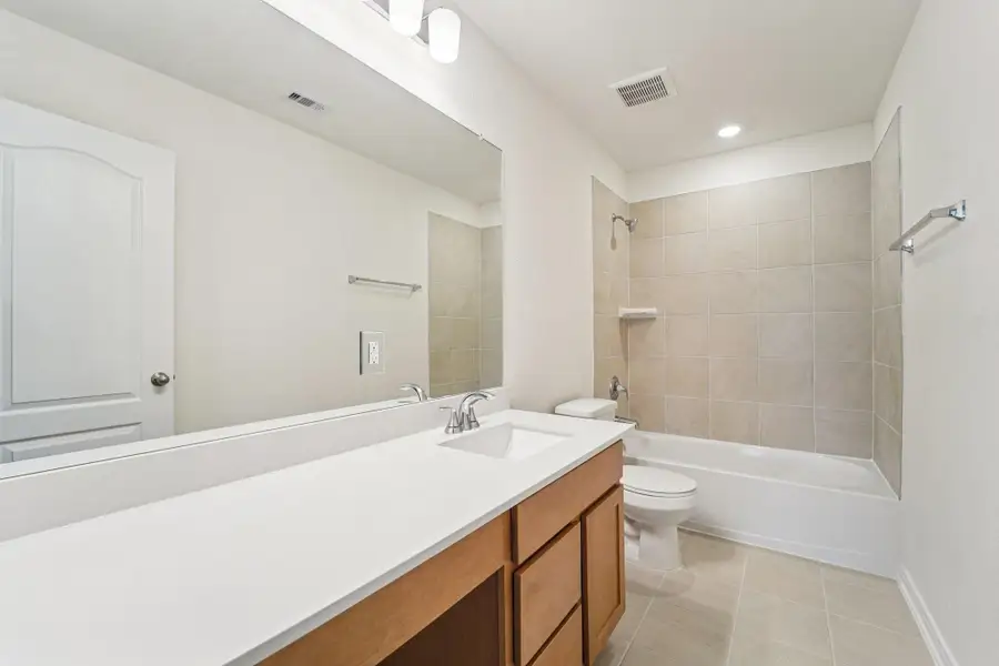 The secondary bathroom offers tile flooring, a bath/shower combination with a stylish tile surround, rich stained wood cabinetry, elegant light countertops, a mirror, and sleek fixtures with modern finishes.