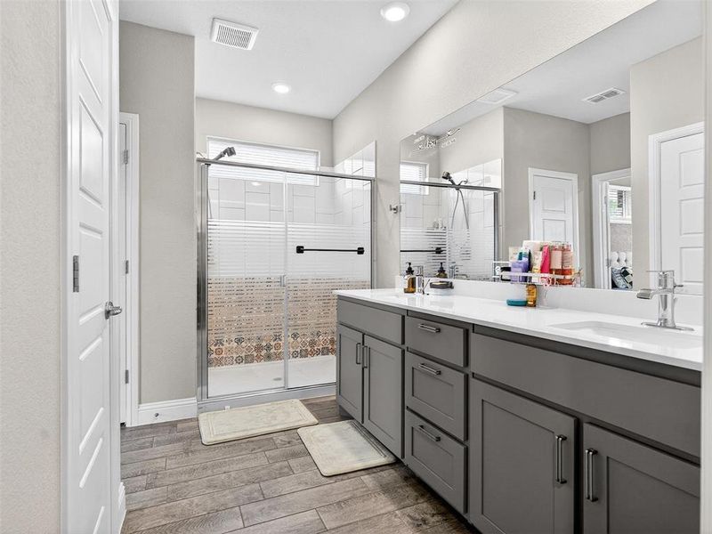 Contemporary bathroom featuring wood-finish flooring, a double vanity with gray cabinetry and white countertops, and a tiled shower with a glass enclosure Contemporary bathroom featuring wood-finish flooring, a double vanity with gray cabinetry and white countertops, and a tiled shower with a glass enclosure