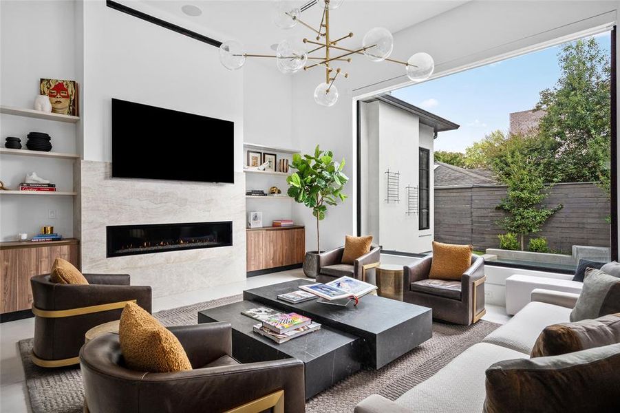 This elegant family room boasts clean lines and modern design elements, including sleek linear air vents, a striking contemporary Perfection Supply gas fireplace, and a stylish brass light fixture suspended from the soaring high ceiling above.