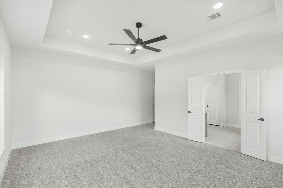 Empty bedroom with tray ceiling, ceiling fan, gray carpet, and open double doors to ensuite bathroom