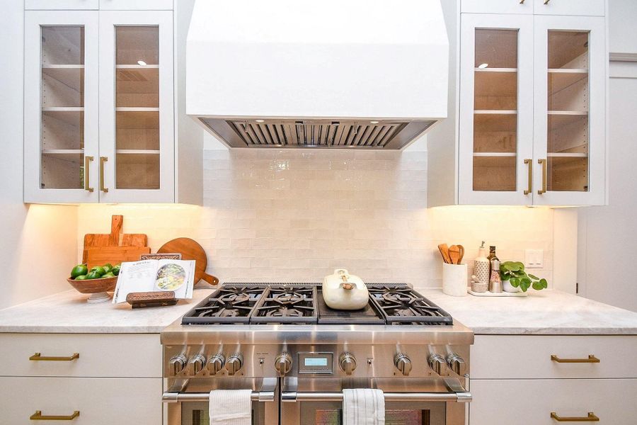 This kitchen features a sleek, modern design with a stainless steel gas stove, white cabinetry with glass doors, and warm under-cabinet lighting. The marble countertop provides a clean and elegant workspace, complemented by stylish brass hardware.