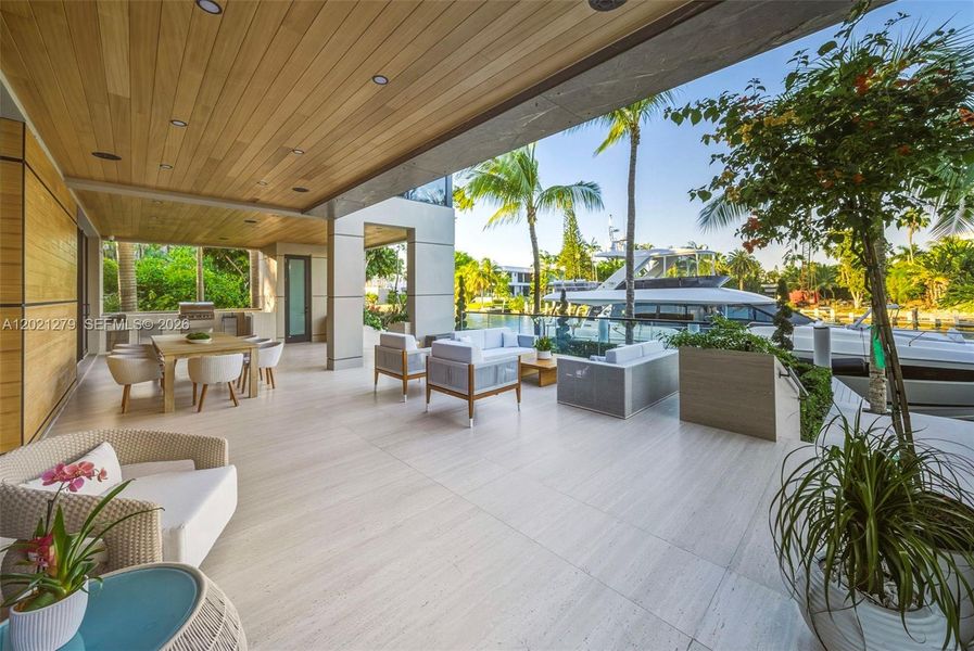 Expansive waterfront ground-level terrace with large-format stone tile, covered loggia with outdoor kitchen and dining, multiple woven lounge seating areas, glass railing, frameless canal views, private dock with large yacht, and tropical landscaping.