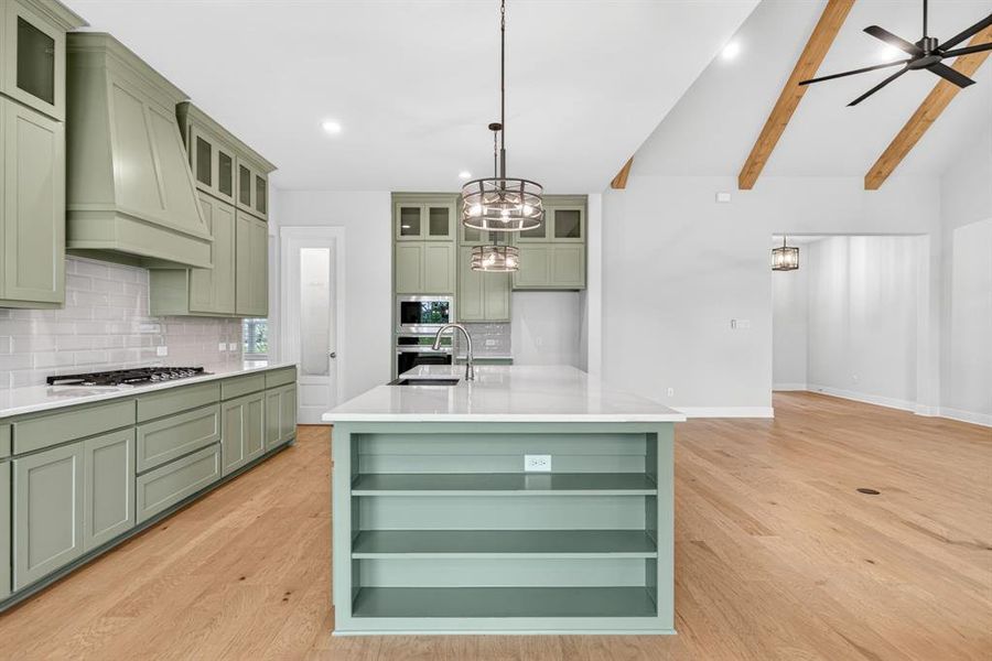 Kitchen and living space featuring a vaulted ceiling with exposed wood beams, a central kitchen island with integrated shelving, light wood-finish flooring, light green cabinetry, and white countertops Kitchen and living space featuring a vaulted ceiling with exposed wood beams, a central kitchen island with integrated shelving, light wood-finish flooring, light green cabinetry, and white countertops