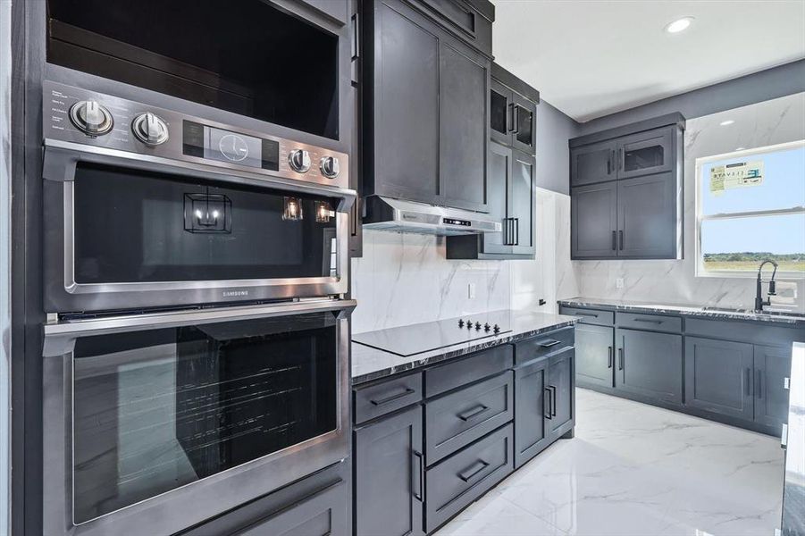 Kitchen featuring dark stone countertops, light marble finish flooring, double oven, tasteful backsplash, and glass insert cabinets