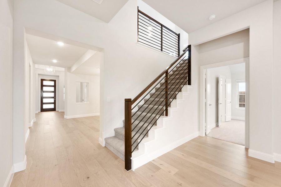 Open foyer with staircase featuring dark wood railings, light hardwood floors, and a glass-panel front door visible in the background.