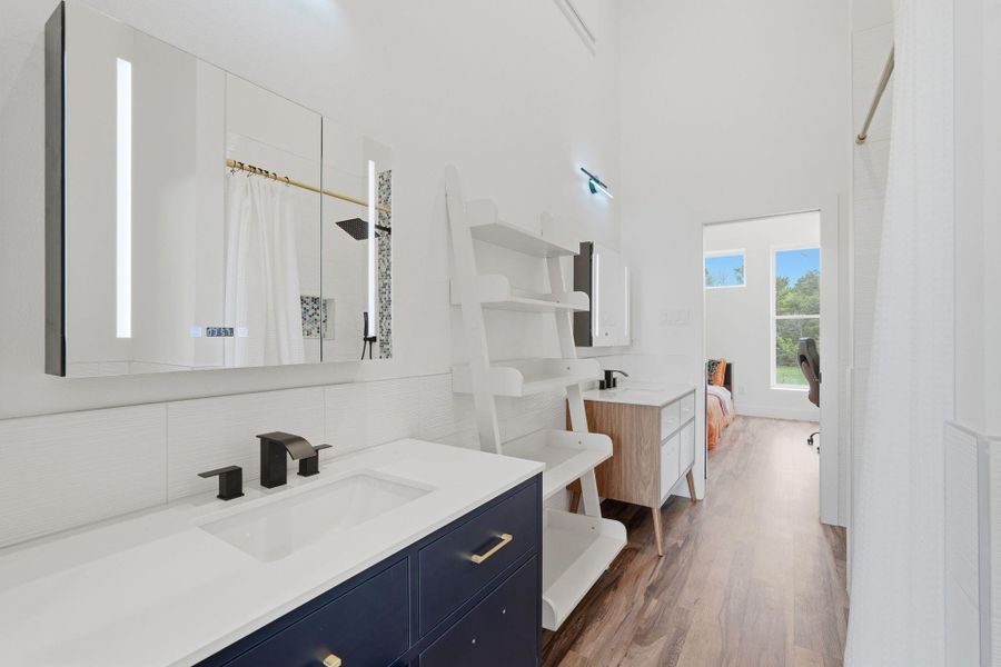 This photo showcases a modern, bright bathroom with a sleek double vanity, contemporary fixtures, and ample storage. The space features a large mirror with built-in lighting and transitions into a bedroom with natural light from large windows.