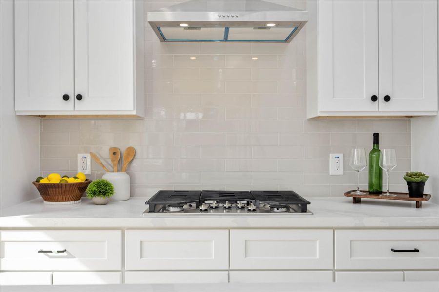 A modern gas cooktop is set against a sleek glossy subway-tile backsplash, framed by bright white cabinetry, polished white quartz countertops, and simple decorative accents that create a clean, contemporary focal point.