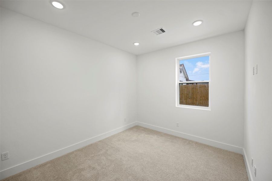 This first level bedroom features a clean, modern design with white walls and recessed lighting. A single window allows natural light, complemented by plush carpeting, creating a cozy and inviting space.