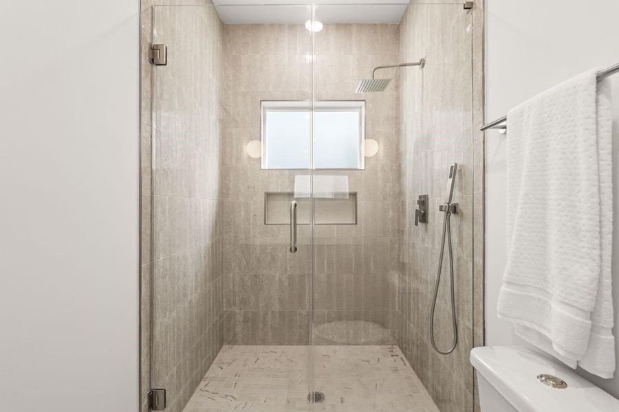 A generous walk-in shower wrapped in floor to ceiling tile creates a spa-like retreat, complete with a rainfall shower head, handheld wand, recessed niche, and frameless glass enclosure.