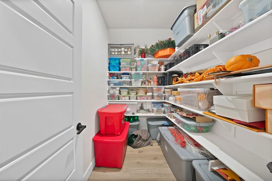 Walk-in storage area featuring extensive built-in shelving, durable wood-finish flooring, a white paneled door, and a matte black door handle Walk-in storage area featuring extensive built-in shelving, durable wood-finish flooring, a white paneled door, and a matte black door handle