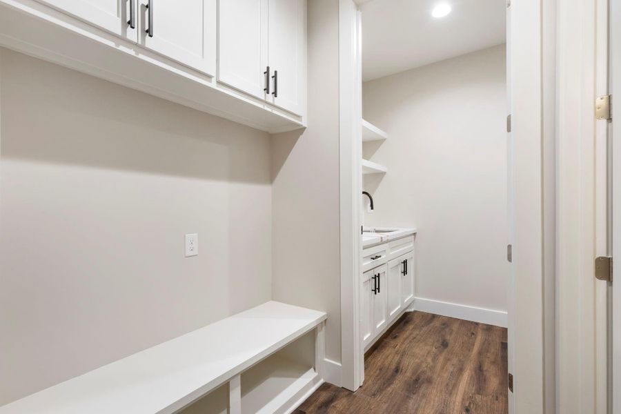 This photo showcases a modern utility or mudroom area with dark wooden floors, white cabinetry, and a built-in bench with upper storage. The adjacent space appears to be a laundry or service room with additional cabinetry and countertop workspace.