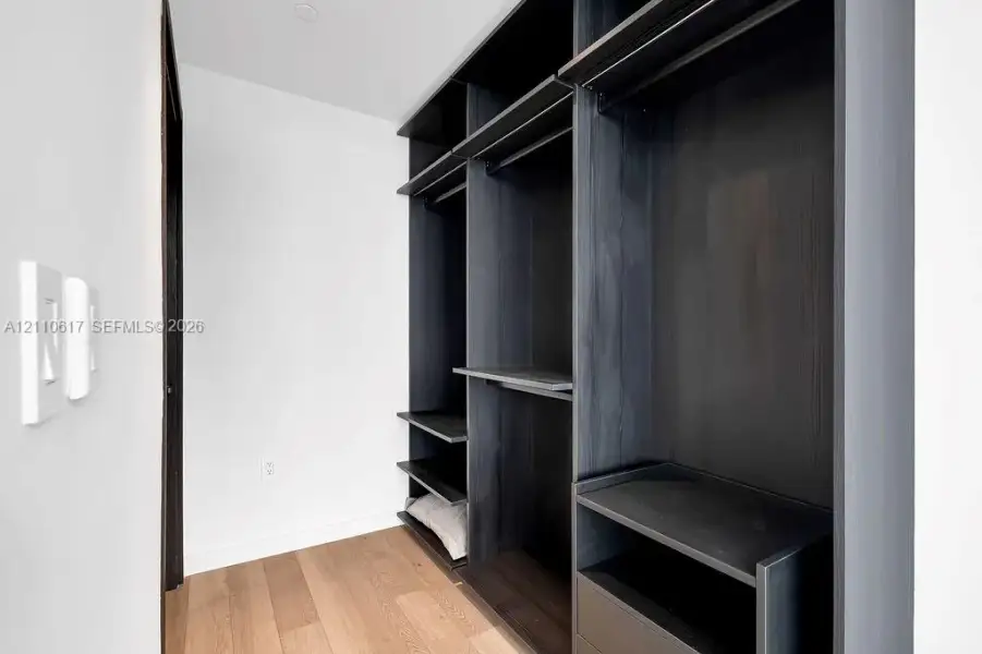 Custom dark-finish built-in cabinetry featuring multi-level shelving, hanging rods, and integrated drawers, paired with light-tone wood-finish flooring Custom dark-finish built-in cabinetry featuring multi-level shelving, hanging rods, and integrated drawers, paired with light-tone wood-finish flooring