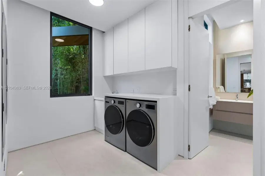 Laundry room featuring floor-to-ceiling cabinetry, custom solid-surface countertops, large-format tile flooring, and a window with natural foliage exposure