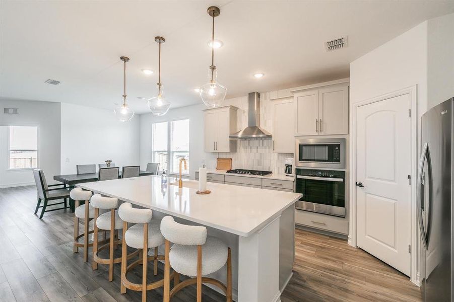 Kitchen with appliances with stainless steel finishes, backsplash, wall chimney exhaust hood, decorative light fixtures, and a center island with sink