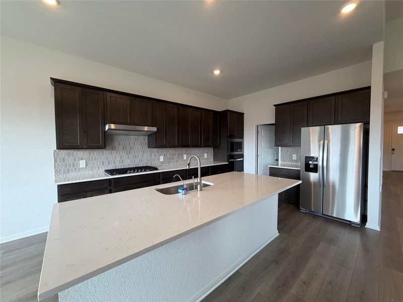 Kitchen featuring tasteful backsplash, stainless steel appliances, dark brown cabinetry, light stone counters, and dark wood-type flooring