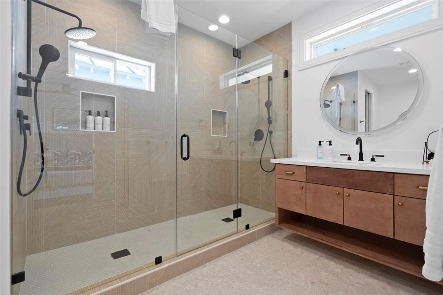 This full bathroom of the primary bedroom blends modern contemporary design with functionality—featuring a spacious enclosed dual-shower setup, black-accented fixtures, a long wood-textured vanity with a sleek black faucet, and an elegant oblong mirror with a soft transparent border. This full bathroom of the primary bedroom blends modern contemporary design with functionality—featuring a spacious enclosed dual-shower setup, black-accented fixtures, a long wood-textured vanity with a sleek black faucet, and an elegant oblong mirror with a soft transparent border.
