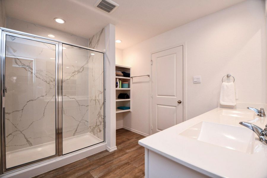 This bathroom includes a glass-enclosed shower with elegant marble-style tiling, a double-sink vanity, and built-in shelving for storage.