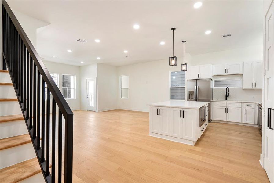 This is a bright, open-concept space featuring a modern kitchen with white cabinetry and stainless steel appliances, an island with a sink, elegant pendant lighting, and hardwood flooring throughout. A staircase with black metal leads to the upper level, and natural light streams in from multiple windows, creating a welcoming atmosphere.