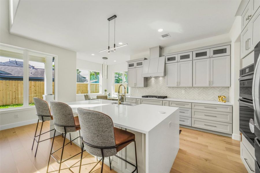 This modern kitchen features sleek white cabinetry, a large island with seating, and stainless steel appliances. It has ample natural light from multiple windows and a view of the backyard, creating a bright and inviting space.