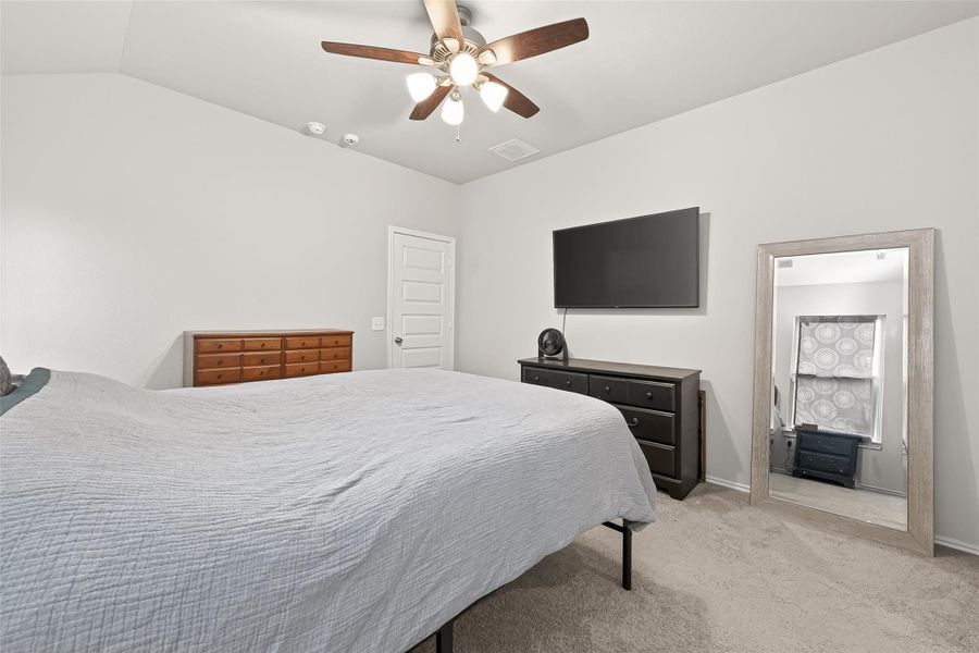 This bedroom is spacious and modern, featuring a ceiling fan, neutral walls, and carpeted flooring. It includes a large bed, two dressers, a wall-mounted TV, and a full-length mirror, creating a comfortable and stylish retreat.