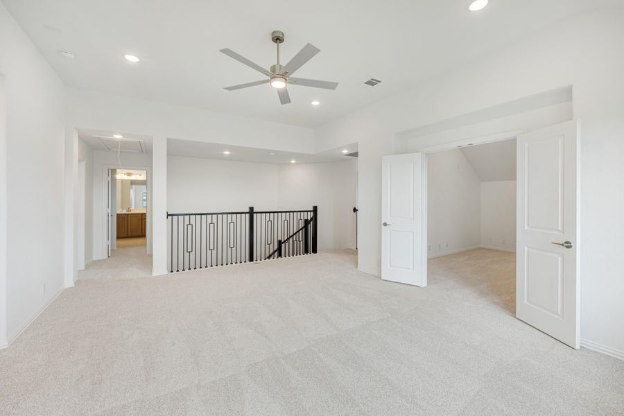Spacious upstairs loft with carpet, ceiling fan, black metal stair railing, and open doors to additional rooms Spacious upstairs loft with carpet, ceiling fan, black metal stair railing, and open doors to additional rooms