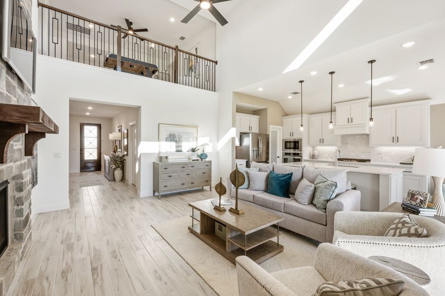Open-concept living room with two-story ceiling, stone fireplace, and kitchen visible in background